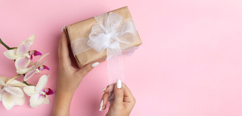 Young female hands hold a gift craft box with a white bow on a pink background with orchid flowers. Holiday concept, Valentine's Day, Women's Day, Thanksgiving, Birthday. Beautiful handmade packaging.