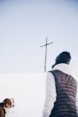 Blick auf dicht verschneites Gipfelkreuz im Winter, im Vordergrund wandernde Frau und Hund die den...