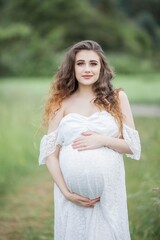 Fototapeta na wymiar A beautiful young pregnant woman with curly long hair in a white dress walks in the field. Spring portrait of a pregnant woman. Happy pregnancy.