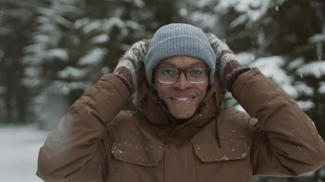 Lockdown portrait of young African man standing in winter forest, taking off hood of the jacket and looking at camera with smile on his face