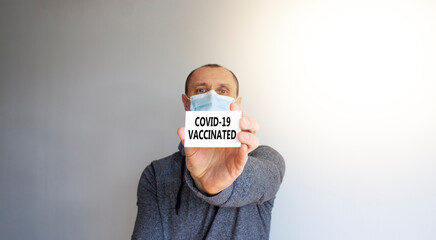 COVID-19 vaccinated symbol. White card with words 'COVID-19 vaccinated'. A young man in a grey wear and medical mask. Sunshine. Beautiful white background. COVID-19 vaccinated coronavirus concept.