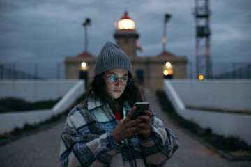 Young woman in winter or autumn outfit look at phone, face lit with screen light. Millennial generation z teenager or young adult attached to smartphone, information overload concept, phone app