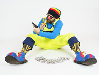 A clown in a bright costume sits and holds a pistol in his hands, which he examines through a magnifying glass