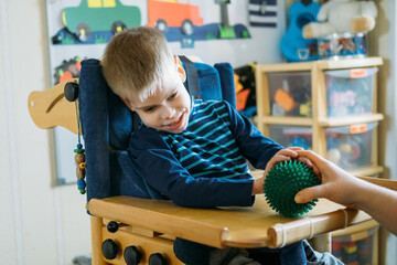 Sensory Activities for kids with disabilities. Preschool Activities for Children with Special Needs. Boy with with Cerebral Palsy in special chair play with mom at home. - obrazy, fototapety, plakaty