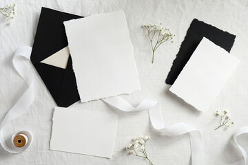 Black envelopes and white paper cards with flowers and ribbon on textile background. Flat lay, top...