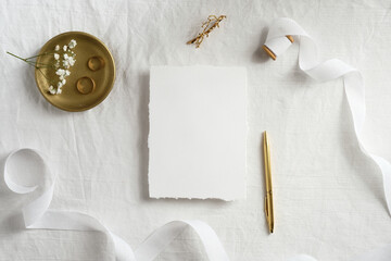 Blank paper card mockup, white ribbon, golden pen and rings on textile background. Top view, flat...