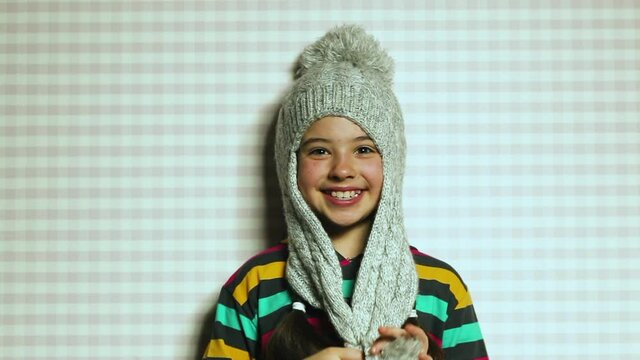 A beautiful dark-skinned little girl in a bright striped sweatshirt and a big winter hat with a bubo makes a grimace. Emotional happy child.