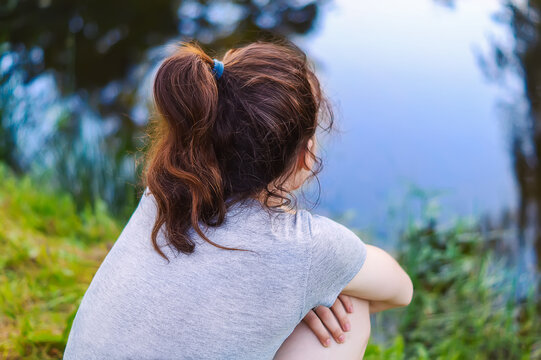 A lonely young girl sits on the bank of a river. A thoughtful Girl, in a gray T-shirt, sits silently near a small lake on the grass and enjoys nature.
