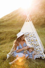 Beautiful mother with her little daughter sitting near wigwam in the field. Spending time together, outside, on vacation, outdoors. Beautiful sunset light in the garden or in the park. © Andriy Medvediuk