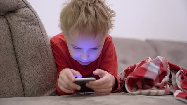 Portrait of smiling cute child lies on sofa and plays cheerfully in a game off phone. Gadget addiction. Mobile addict concept. Surfing the Internet.