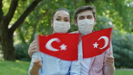 Two young people are waving the Turkish flag while looking at the camera. They wear a medical mask...