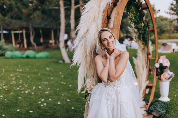 A beautiful blonde bride in a long dress stands near a wooden wedding arch decorated with reeds,...