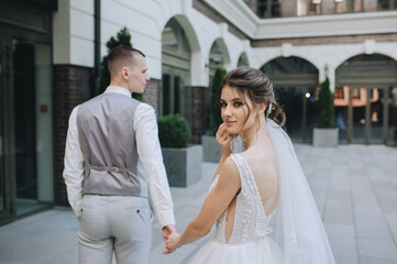 Fototapeta premium The brunette curly-haired bride turned around in a white dress with a long veil smiling, looking into the frame, walking in the city, holding the groom's hand. Wedding portrait of the newlyweds.
