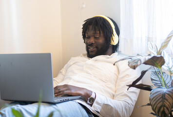 Smiling afro man works on a sofa at home with a laptop and headphones