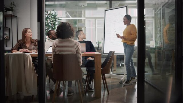 Project Manager Makes a Presentation for a Young Diverse Creative Team in Meeting Room in an Agency. Colleagues Sit Behind Conference Table and Discuss Business Development, User Interface and Design.