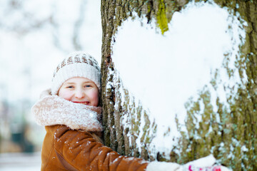 smiling stylish girl hugging tree with snowy heart