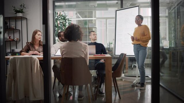 Project Manager Makes a Presentation for a Young Diverse Creative Team in Meeting Room in an Agency. Colleagues Sit Behind Conference Table and Discuss Business Development, User Interface and Design.