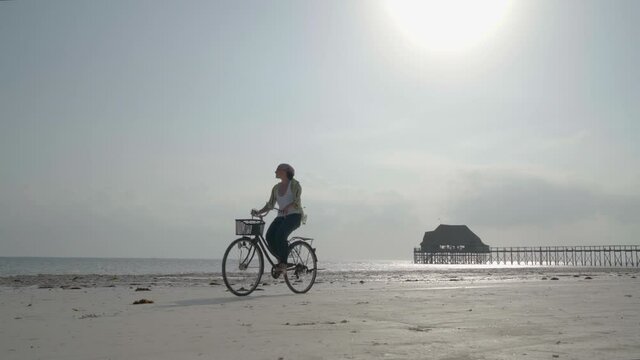 4K footage of a female dressed light summer clothes riding old vintage bicycle with front basket on the lonely low tide ocean white sand coast on Kiwengwa beach on Zanzibar island, Tanzania.