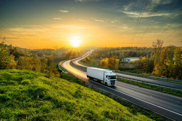 White truck driving on the highway winding through forested landscape in autumn colors at sunset - obrazy, fototapety, plakaty