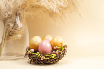 Pampas grass in glass vase, yellow and pink painted eggs in nest on beige background. Easter holiday, modern minimal composition. Eco Stylish decor concept.