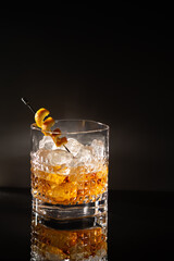 Elegant chrystal glass of Whiskey with ice cubes and orange peel spiral garnish on black with reflection.Close-up of noble acoholic drink.