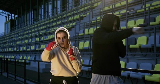 Young boxing boy trainer and novice girl train boxing punches in boxing gloves on the sports field at night