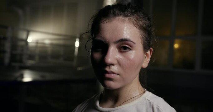 Sweaty female fighter during break in gym. Tired young woman with sweaty face resting during break in boxing workout in dark gym
