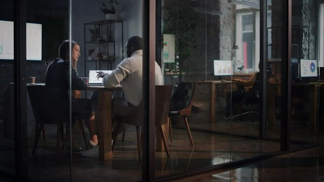Two Diverse Multiethnic Colleagues Have a Conversation in a Meeting Room Behind Glass Walls in an Agency. Female Creative Director and African American Project Manager Discuss Work on Laptop Computer.