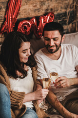 Young smiling happy couple in love in comfortable home clothing clinking glasses owith sparkling wine and celebrating holiday at home. Valentines day celebration concept