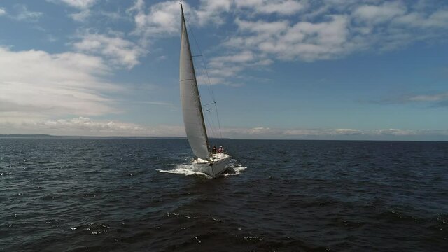 Aerial view. A white sailboat is moving fast on the waves of the sea. A boat in full sailing gear floats on camera. Close hauled sailing in ocean. Yacht in windy and sunny day. Drone view.