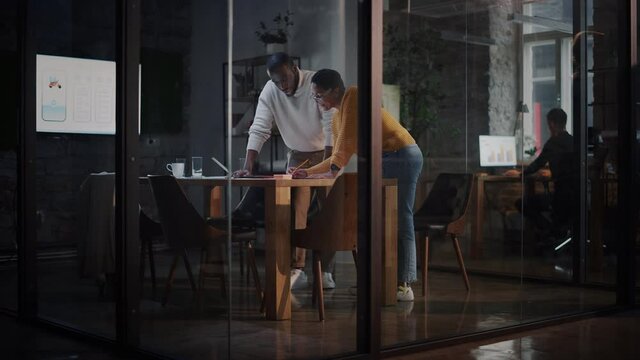 Two Diverse Multiethnic Colleagues Have a Conversation in a Meeting Room Behind Glass Walls in an Agency. African American Creative Director and Female Project Manager Discuss Work on Laptop Computer.