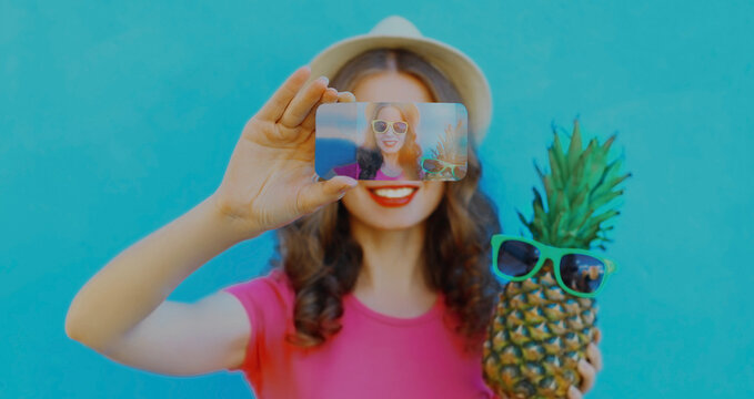 Summer image of woman stretching her hands taking selfie picture by phone with pineapple wearing a sunglasses on a blue background