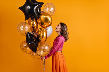 Debonair girl looking at party balloons. Indoor photo of pleased woman in pink blouse isolated on orange background.