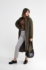 Young attractive woman in long green coat with black handbag on white background. Model in gray short pants, brown sweater and black shoes looking to the right