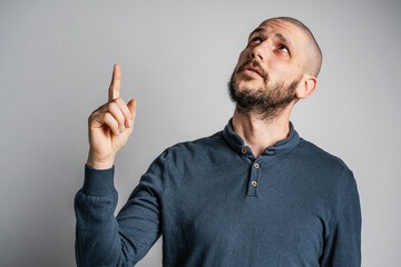 Front view portrait of adult caucasian man with beard and short hair pointing up while standing in front of white wall thinking with copy space studio shot