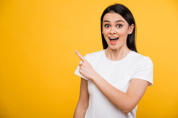 Excited shocked caucasian beautiful brunette woman in a white t-shirt, points finger to the side at blank space for text or advertisement, while standing against isolated orange background