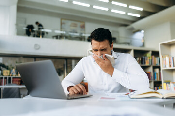 Fototapeta na wymiar Hispanic business man sneezing, using a paper tissue, feeling unwell. Unhealthy manager struggling with running nose, catch a flu, remotely working or studying, while illness