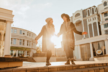 Elegant girls holding hands in sunny october day. Outdoor portrait of european ladies walking around city.