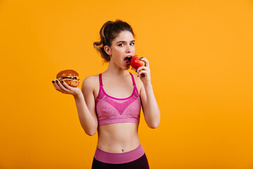 Sporty woman eats red apple on orange background. Relaxed girl holding tasty hamburger.
