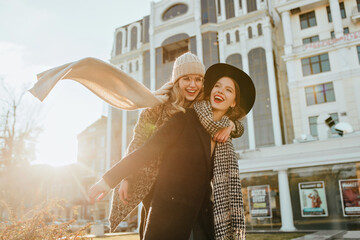 Pretty girl in long scarf enjoying weekend with friend. Outdoor photo of two joyful ladies fooling around in autumn.