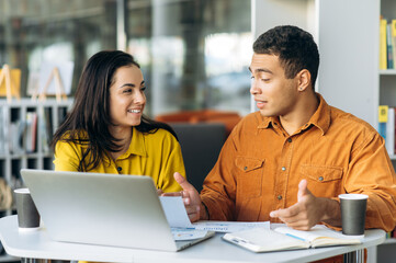 Two joyful multiracial colleagues or students, caucasian girl and hispanic guy, are using a laptop while sitting at workplace discussing about job or education and smiling
