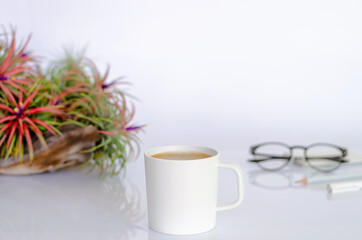 A cup of coffee on working table with air plant Tillandsia, spectacles, pen and pencil on white background.