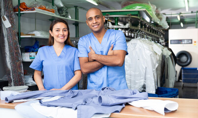 Naklejka premium Smiling female worker of modern laundry standing with her colleague at reception counter, showing clean clothing and offering professional dry cleaning