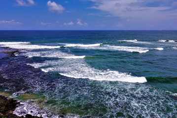 On the turquoise surface of the Indian Ocean, white lace foam of waves. There are picturesque clouds in the azure sky. Summer sunny day in Sri Lanka