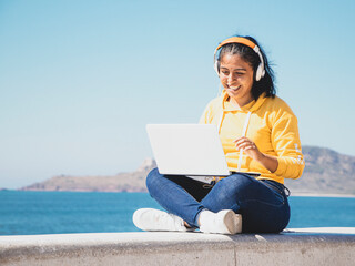 Foto mujer joven latina sentada junto al mar sonriendo escuchando música con pantalón y camisa...