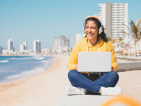 foto mujer estudiante escuchando m&uacute;sica con aud&iacute;fonos en laptop junto al mar vestida de amarillo