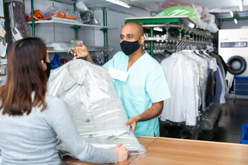 Positive man laundry worker in protective mask for disease prevention returning clean clothes to...