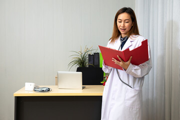 Young Asian Smiling female doctor holding and checking red medical records file in Examination room