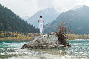 A man stands on a stone in the center of a mountain lake and practices yoga. Pose Vrikshasana.