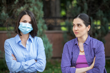 Two young women sits on a summer park bench. One woman wearing a mask, one without a mask....